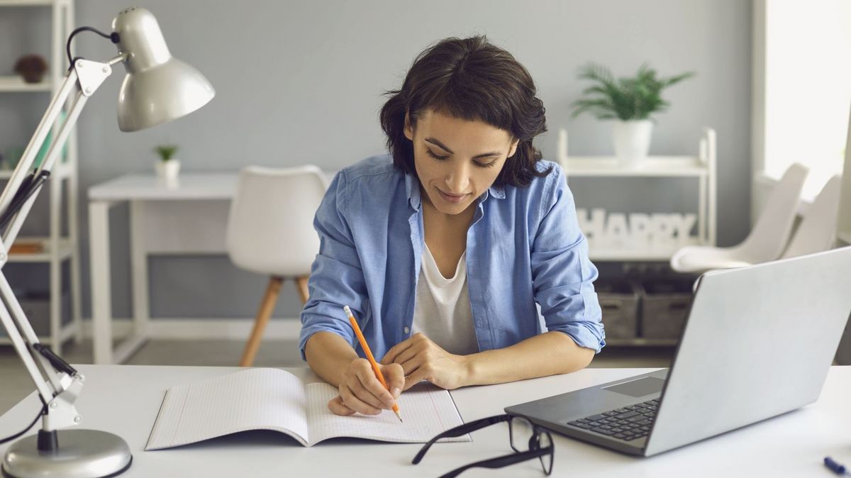 Frau in einem Wohnzimmer an einem Tisch, auf dem ein Laptop steht, die Frau schreibt in ein Heft.