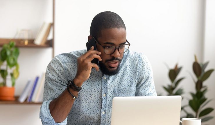 Mann in einer Küche telefoniert mit einem Smartphone und blickt in einen Laptop