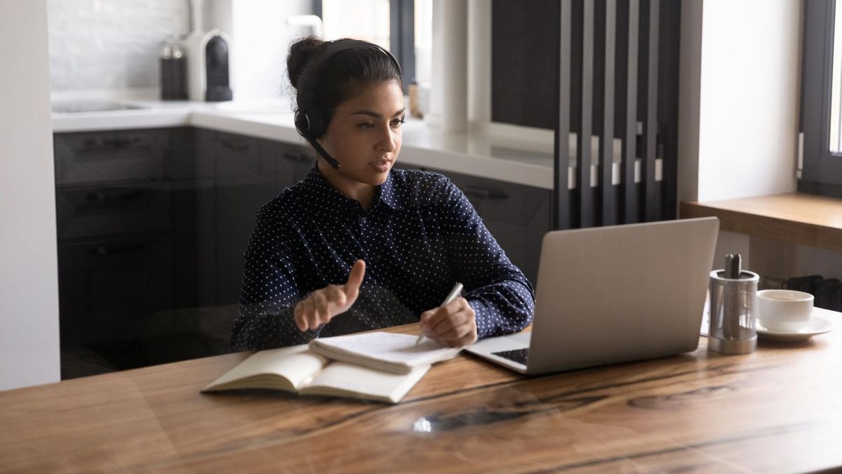 Junge Frau mit Head-Set am Schreibtisch, auf dem ein Laptop steht. Sie spricht.