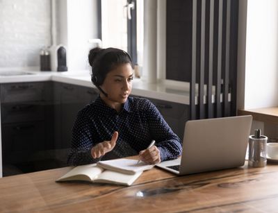 Junge Frau mit Head-Set am Schreibtisch, auf dem ein Laptop steht. Sie spricht.